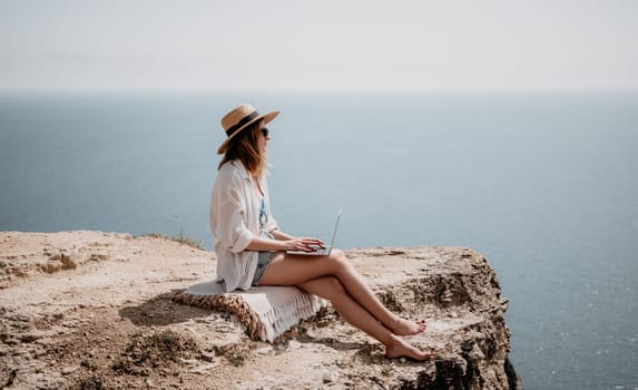 Successful business woman in yellow hat working on laptop by the sea. Pretty lady typing on computer at summer day outdoors. Freelance, travel and holidays concept.