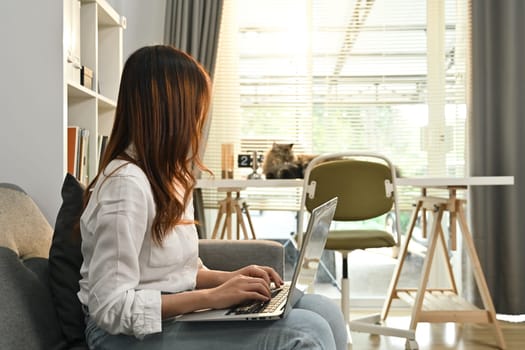 Side view of young woman relaxing on couch and checking email or online news on laptop. People, technology and lifestyle concept.