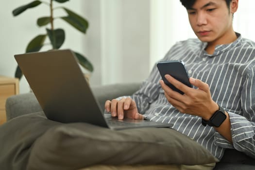 Millennial asian man sitting on couch with laptop and using smart phone. People, technology and lifestyle concept.