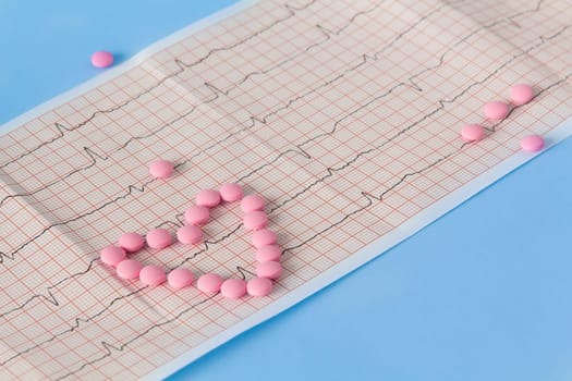 A large handful of pink pills lie in the form of a heart on an electrocardiogram, on a blue background. The concept of a healthy lifestyle and timely medical examination