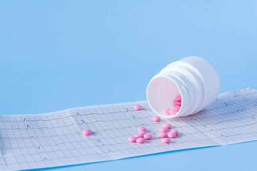 A large handful of pink pills poured out of a white jar on an electrocardiogram of the heart, on a blue background. The concept of a healthy lifestyle and timely medical examination