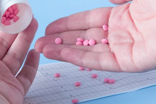 A hand pours a handful of pink pills from a white jar onto an electrocardiogram of the heart, on a blue background. The concept of a healthy lifestyle and timely medical examination