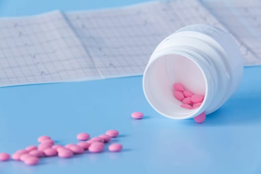 A large handful of pink pills poured out of a white jar on an electrocardiogram of the heart, on a blue background. The concept of a healthy lifestyle and timely medical examination