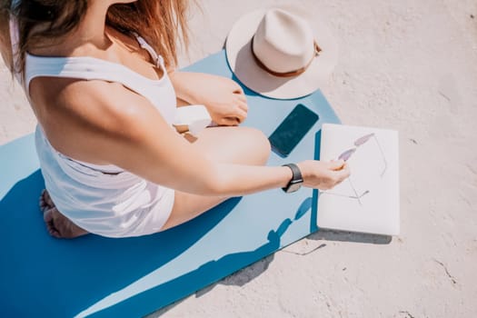 Successful business woman in yellow hat working on laptop by the sea. Pretty lady typing on computer at summer day outdoors. Freelance, travel and holidays concept.