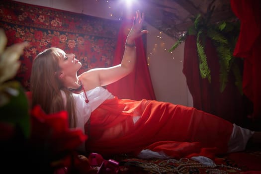 Beautiful European girl looking like Arab woman in red room with rich fabrics and carpets in sultan harem. Photo shoot of an oriental style odalisque. A model poses in sari as indian woman in india