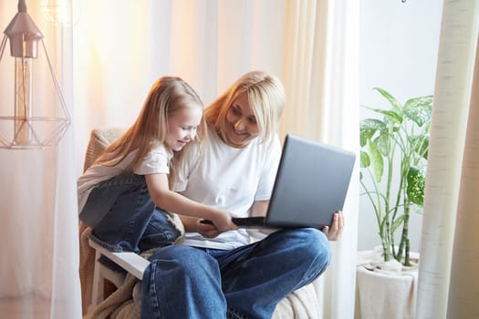 Beautiful young woman and her little cute daughter are using laptop at home. Enjoying spending time together with Internet and modern technologies