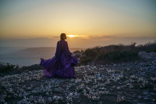 Sunset purple dress woman mountains. Rise of the mystic. sunset over the clouds with a girl in a long purple dress. In the meadow there is a grass dream with purple flowers