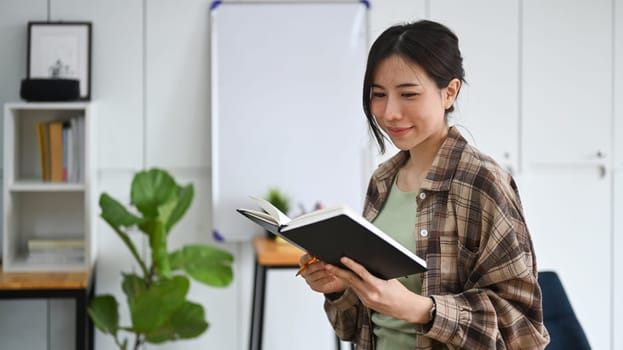 Smiling woman standing in office and reading a book.