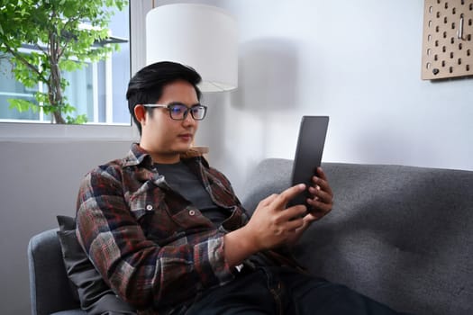 Young man sitting on sofa and browsing internet with digital tablet.