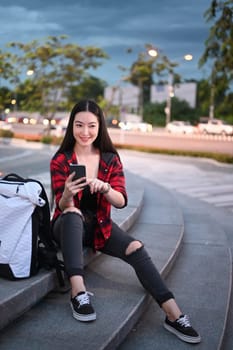 Full length portrait of smiling young woman sitting on stairs in the city and using smart phone.