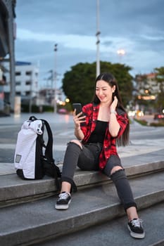 Portrait of smiling young woman sitting on stairs in the city at night and using smart phone.