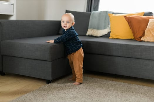 Toddler boy laughing having fun standing near sofa in living room at home. Adorable baby making first steps alone. Happy childhood and child care
