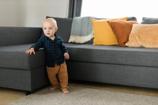 Toddler boy laughing having fun standing near sofa in living room at home. Adorable baby making first steps alone. Happy childhood and child care