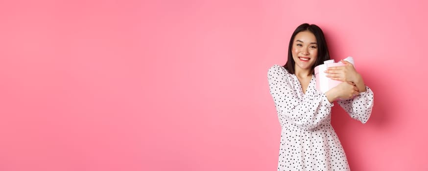Lovely asian woman hugging her gift and smiling thankful, receive valentines day present, standing over pink background.