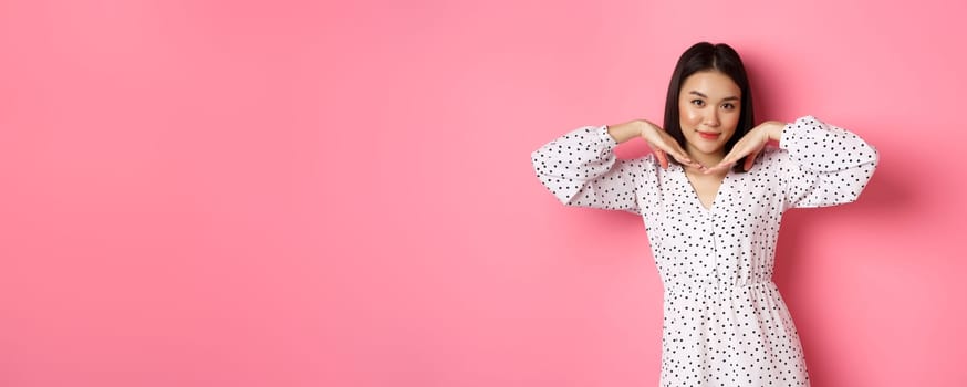Beautiful asian woman in trendy dress making cute face, holding hands near jawline and gazing coquettish at camera, standing over pink background.