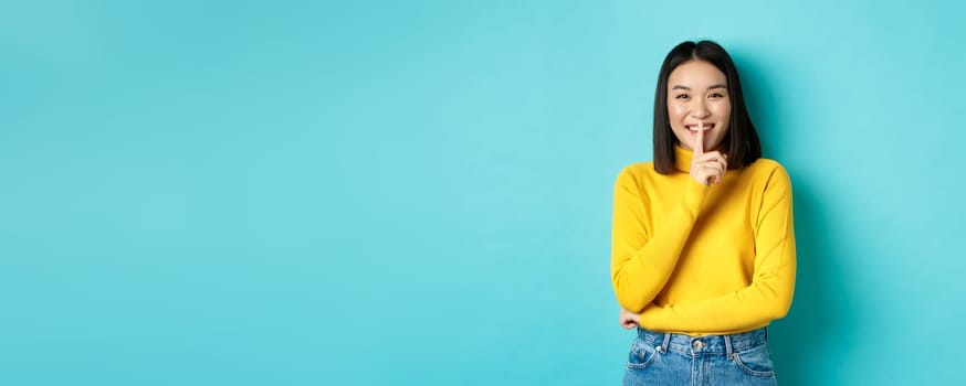 Cheerful asian girl asking to be queit, shushing at camera and smiling, keeping secret, standing against blue background.