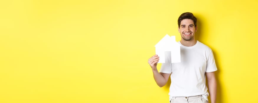 Real estate concept. Young man in white t-shirt holding paper house model and smiling, searching for apartment, standing over yellow background.