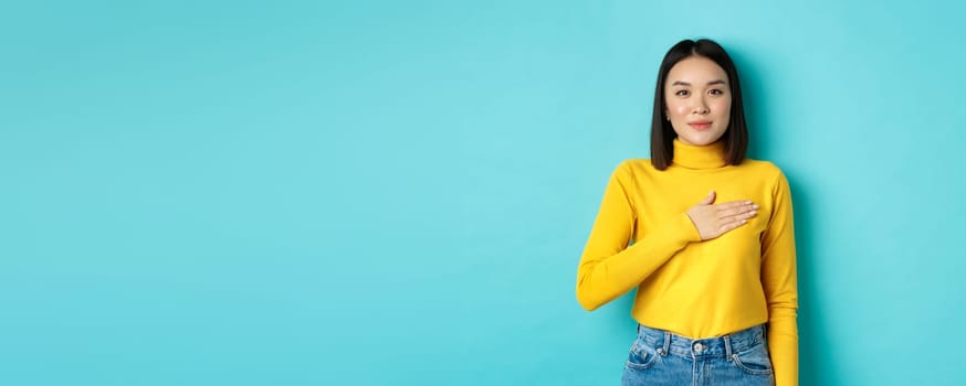 Image of proud smiling asian woman holding hand on heart, showing respect to national anthem, standing over blue background.