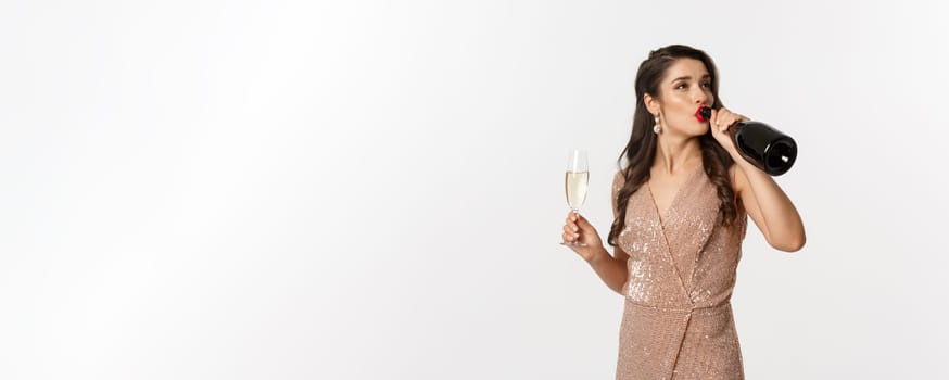 Winter holidays celebration concept. Careless woman in elegant dress drinking champagne from bottle and looking unbothered, standing over white background.