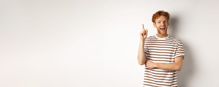 Young man with red hair having an idea, raising finger to say suggestion, standing happy over white background.
