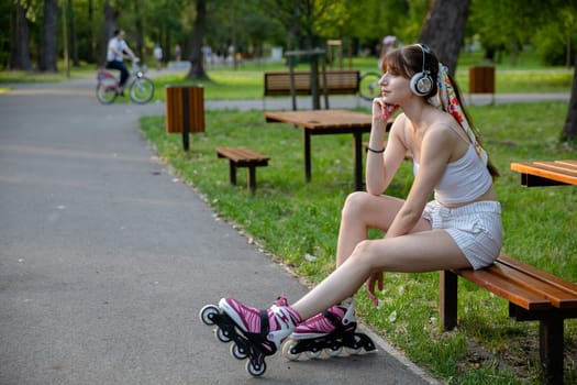 A thoughtful woman looks ahead while sitting on a bench. She supports her head with her hand. The woman is wearing wireless headphones on her head. In the background you can see a blurry figure riding a bicycle.