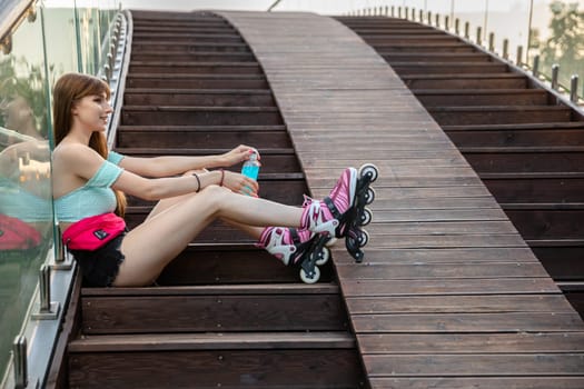 A girl in a summer outfit sits on the steps. She opens a bottle of blue isotonic drink. The brunette with long hair is wearing purple rollerblades