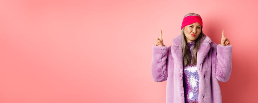 Disappointed stylish asian lady pointing fingers up. Senior woman in trendy fake fur coat grimacing skeptical and pointing fingers up, dislike promo, standing over pink background.