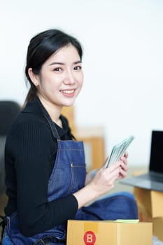 Starting small business entrepreneur of independent Asian woman smiling and holding money using laptop computer with cheerful success of online marketing package box items and SME delivery concept.