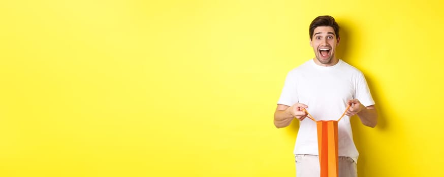 Surprised guy open shopping bag with fist, looking excited and happy at camera, standing against yellow background.