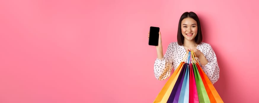 Attractive asian woman showing smartphone app and shopping bags, buying online via application, standing over pink background.