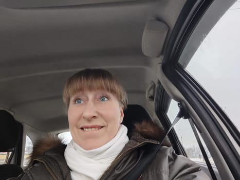 Portrait of adult cute funny blonde woman in warm clothes taking a selfie inside the car and driving it. The concept of pleasant trip in spring, autumn, winter