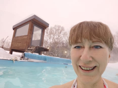 Adult mature woman takes selfie in a pool with warm hot termal mineral water in winter and white snow around. Wellness center and the concept of health care. Travel, recreation, medicine and rest