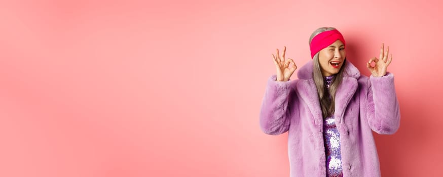 Fashion and shopping concept. Stylish asian granny saying all okay and winking. Trendy senior woman in winter fake-fur coat show OK sign, praising good product, pink background.