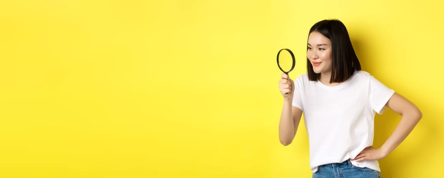 Young asian woman searching for something, looking left through magnifying glass and smiling pleased, investigating, standing over yellow background.