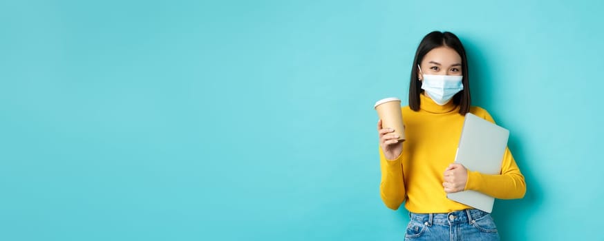 Covid, pandemic and social distancing concept. Stylish asian woman wearing medical mask, holding cup of coffee and laptop, going to work, standing over blue background.