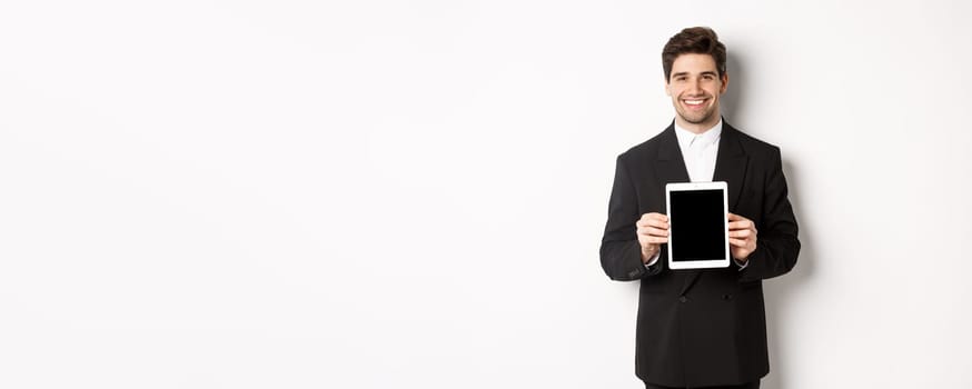 Portrait of handsome businessman in trendy suit, showing digital tablet screen and smiling, standing against white background.