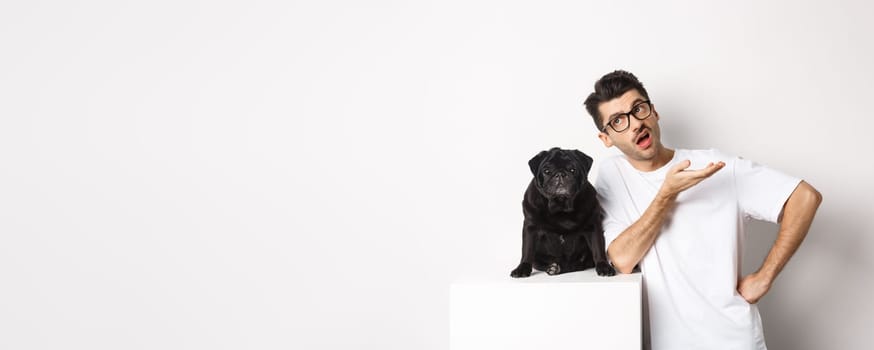 Image of hipster guy talking with his dog, looking skeptical and gesturing, having conversation with cute black pug, standing over white background.