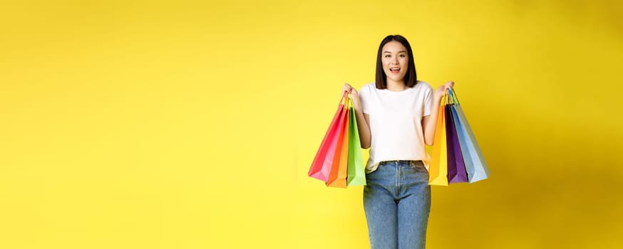 Full size portrait of beautiful asian girl going shopping, holding paper bags from stores and smiling, standing in jeans and white t-shirt over yellow background.