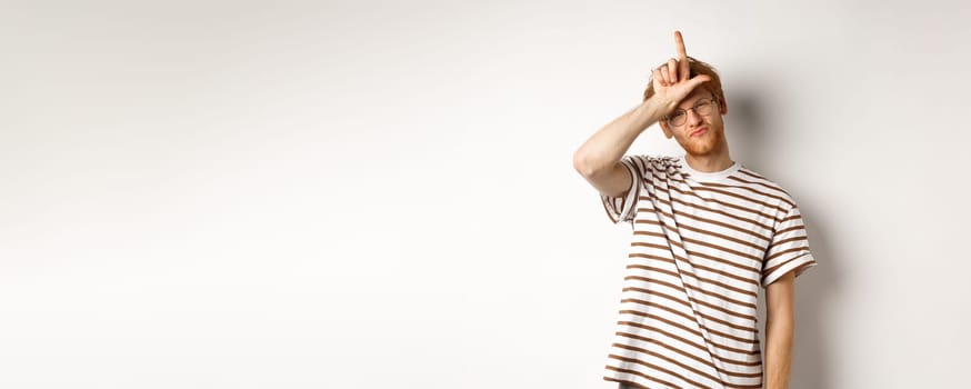 Sassy and arrogant redhead man scolding lost team, showing loser gesture on forehead and making smug face, standing over white background.