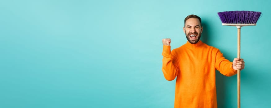 Image of encouraged and motivated man getting ready for cleaning, holding broom and making fist pump gesture, standing over turquoise background.