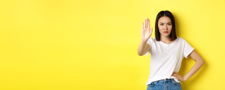Confident and serious asian woman tell no, showing stop gesture to prohibit and warn, disagree with someone, standing upset against yellow background.