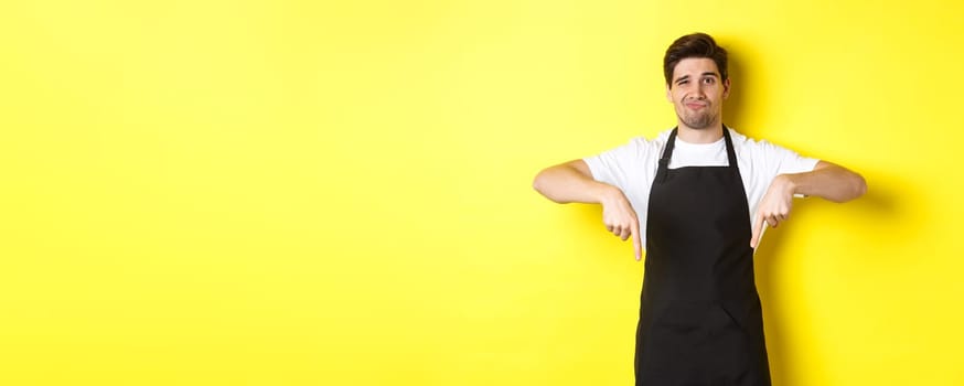 Skeptical barista in black apron pointing fingers down at bad product, looking displeased and unamused, standing over yellow background.