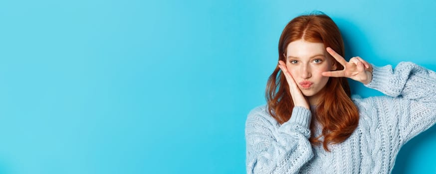 Close-up of beautiful and sassy girl with red hair, showing peace kawaii sign and gazing at camera, standing over blue background.