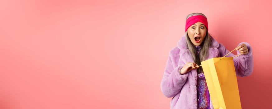 Shopping and fashion concept. Surprised asian grandmother open shop bag and looking amazed at camera, receive mother day gift, pink background.