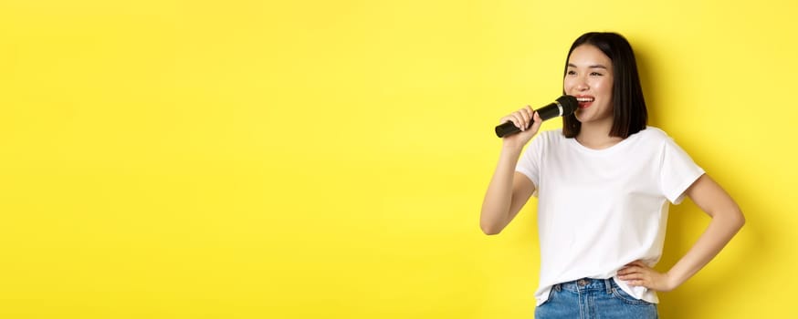 Happy asian woman singing song in karaoke, holding microphone and looking aside with cheerful smile, standing over yellow background.