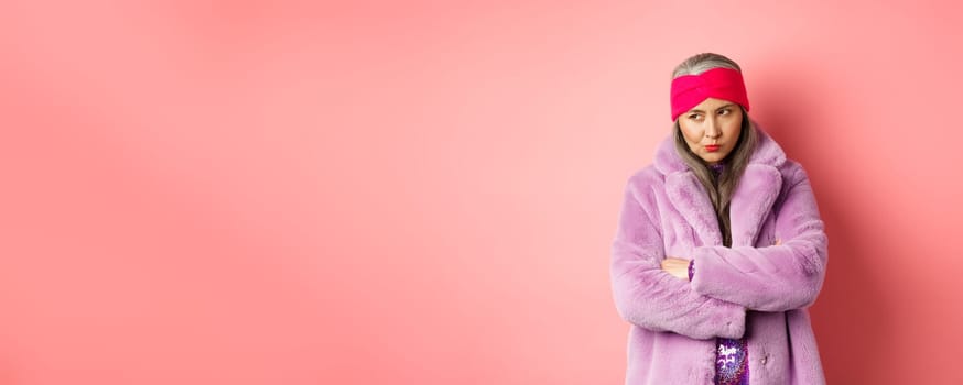 Grumpy funky old asian lady looking from under forehead, pouting and looking left with angry and disappointed face, standing in stylish purple faux fur coat against pink background.