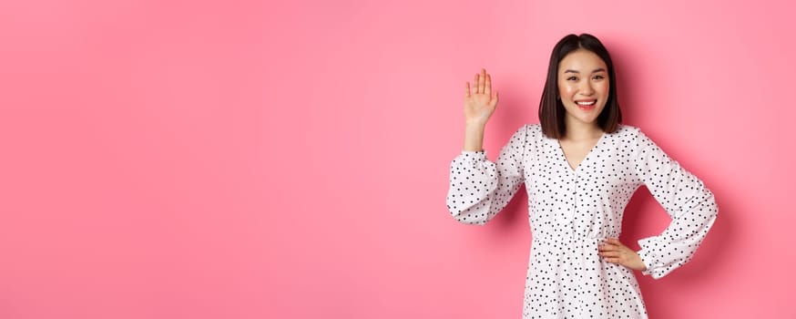 Beautiful asian woman in dress saying hello, waving hand to greet and say hi, smiling friendly at camera, standing over pink background.