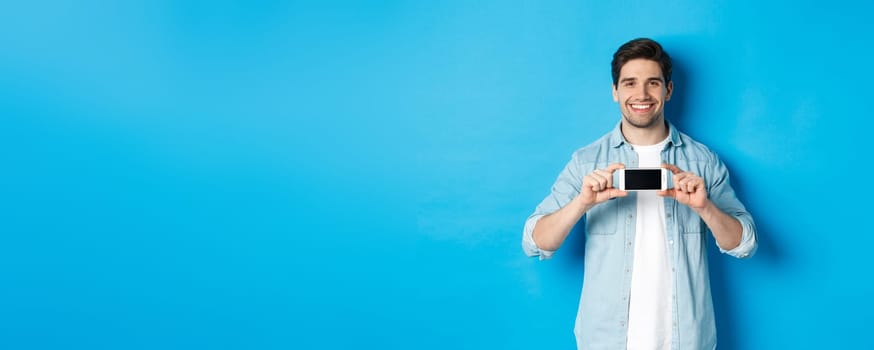 Handsome smiling man showing smartphone screen, standing against blue background for copy space.