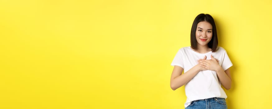 Beauty and fashion concept. Heartfelt asian girl holding hands on heart and smiling touched, thanking you, standing over yellow background.