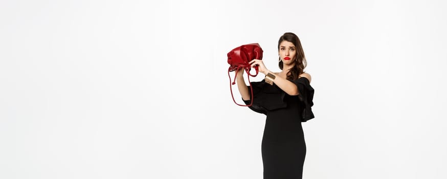 Beauty and fashion concept. Full length of sad young woman in black dress and high heels showing empty purse, sulking disappointed, standing over white background.